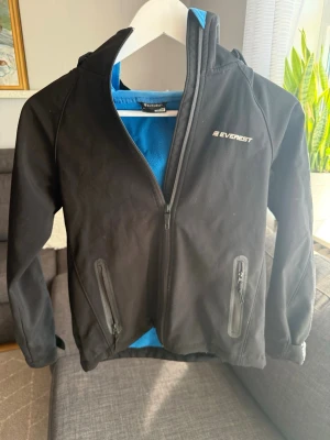 Svart vindjacka från Everest - Snygg svart vindjacka från Everest med blått fleecefoder. Jackan har två dragkedjeförsedda fickor framtill och hög krage. Perfekt för blåsiga dagar och enkel att matcha med olika outfits.