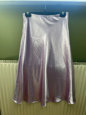 Ljuslila satinkjol med dragkedja - Säljer en glansig ljuslila långkjol i satinliknande material. Kjolen har en enkel och stilren design med en diskret dragkedja i sidan. Perfekt för dig som vill ha en flowy och elegant look.