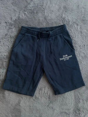 Mörkblå shorts Peak Performance - Snygga mörkblå shorts från Peak Performance med resårmidja och snörning. Shortsen har två sidofickor och logga tryckt på vänster ben. Perfekta för sommaren och tillverkade i mjuk bomull för maximal komfort.