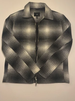 Gråsvart overshirt från H&M - Overshirt från H&M i gråsvart rutigt mönster med dragkedja framtill. Klassisk krage och raka ärmar. Materialet är mjukt och borstat, vilket ger en skön känsla och trendig look. Perfekt att bära över en t-shirt för en avslappnad stil.