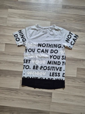 Ljusgrå t-shirt med svart textprint - Snygg ljusgrå t-shirt med svart textprint över hela framsidan och ärmarna. T-shirten har en svart kant längst ner och är tillverkad i mjuk bomull. Passformen är normal och modellen har korta ärmar. Perfekt för dig som gillar statement-plagg.