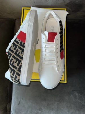 Fendi vita sneakers med FF-mönster - Snygga vita sneakers från Fendi med ikoniskt FF-mönster i svart och beige på sidorna och röda detaljer vid snörningen. Skorna har en klassisk låg siluett, vita snören och en vit sula. Perfekta för dig som vill sticka ut med exklusiv design.