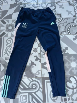 Ajax träningsbyxor från Adidas - Mörkblå träningsbyxor från Adidas med Ajax-logga och 3 stjärnor på vänster ben. Byxorna har ljusrosa och ljusblå detaljer längs benen samt elastisk midja. Tillverkade i ett mjukt och stretchigt material som passar perfekt till träning eller chill.passar till unga mellan 15-16 upp till 176 cm långa 