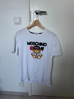 Vit Moschino t-shirt med björntryck - Säljer en vit t-shirt från Moschino med ett stort tryck av en söt björn och Moschino-loggan på bröstet. T-shirten har rund halsringning och korta ärmar. Perfekt för dig som vill ha en statement tee från ett känt märke. Used but still in cood condition. Size M