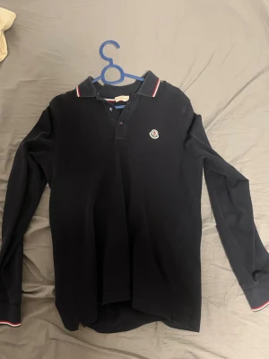 Svart  Moncler pikétröja - Snygg svart långärmad pikétröja från Moncler med klassisk krage och knappar framtill. Tröjan har Monclers ikoniska logotyp på bröstet och diskreta rödvita detaljer på kragen och ärmsluten. Tillverkad i bomull för en bekväm känsla. Storlek S och sitter lite tajt och löst samtidigt så normal passform. Äkta!!! 