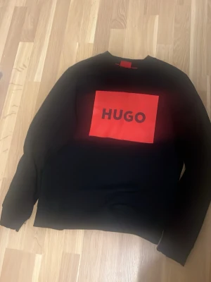 Svart sweatshirt från HUGO BOSS - Svart sweatshirt från HUGO med stor röd logga på bröstet. Tröjan har en klassisk rund halsringning och ribbade muddar vid ärmslut och nederkant. Tillverkad i mjukt material som känns skönt mot huden. Perfekt för en stilren och trendig look.
