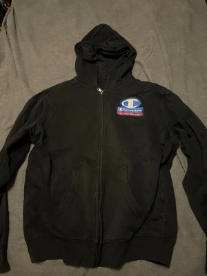 Svart zip-hoodie från Champion - Svart hoodie med dragkedja från Champion. Stor logga på bröstet i blått, vitt och rött. Klassisk huva och två fickor framtill. Perfekt för en avslappnad och sportig stil.