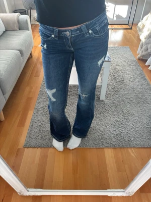 Blå bootcut jeans med slitningar - Säljer ett par blå bootcut jeans med snygga slitningar och detaljer på benen. Jeansen har fem fickor, låg midja och klassisk knappgylf. Perfekta för en avslappnad och trendig look. Använda 2 ggr. Gina 14+ kollektionen. 