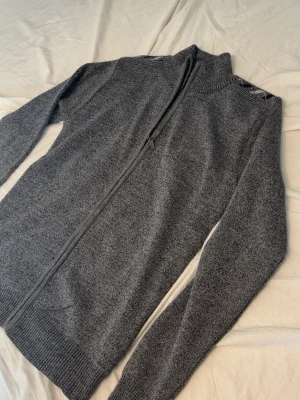 Helzip - Mysig mörkgrå stickad kofta med hel dragkedja framtill och hög krage. Koftan har långa ärmar och ribbade muddar vid ärmslut och nederkant. Perfekt för lager på lager och enkel att matcha med olika outfits.