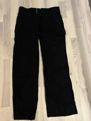 Svarta vida byxor med fickor - Säljer ett par svarta byxor med raka, vida ben och flera fickor. Byxorna har en enkel design utan synliga loggor eller detaljer och är tillverkade i ett mjukt tyg som ser bekvämt ut. Perfekta för en avslappnad och stilren look.