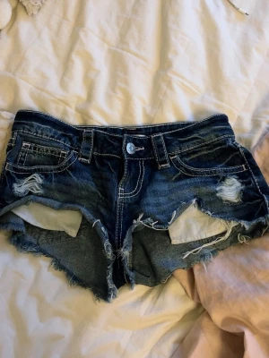 Mörkblå slitna jeansshorts - Säljer ett par mörkblå jeansshorts med slitna detaljer och råa kanter. Shortsen har fem fickor, bälteshällor och dekorativa nitar på bakfickorna. Perfekta för sommaren och ger en avslappnad streetstyle-look.