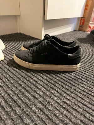 Svarta sneakers från Axel Arigato - Snygga svarta sneakers från Axel Arigato med klassisk låg profil och vit sula. Skorna har ovandel i mocka och textil, svarta skosnören och diskret logga på sidan. Perfekta för en clean och stilren look.