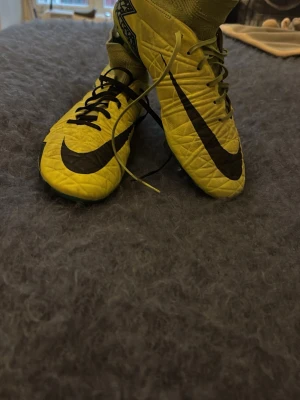 Gula Nike fotbollsskor med strumpa - Säljer ett par gula Nike fotbollsskor med svart swoosh och högt strumpskaft för extra stöd. Skorna har snörning och är tillverkade i syntetmaterial med mönstrad ovandel för bättre bollkontroll. Perfekta för dig som vill sticka ut på planen.
