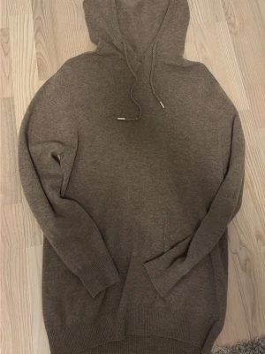 Brun hoodie i mjuk stickad kvalitet - Säljer en  brun hoodie i mjuk stickad kvalitet. Hoodien har huva med dragsko, långa ärmar och ribbade muddar vid ärmslut och nederkant. Perfekt för en chill och avslappnad stil. Har en defekt vid halsen