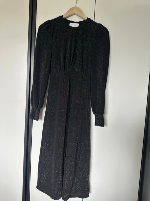 Svart glittrig långklänning Sisters Point - Svart långklänning från Sisters Point med glittrigt tyg och långa puffärmar. Klänningen har rund halsringning och breda manschetter vid ärmsluten. Perfekt för dig som vill sticka ut med en elegant look.