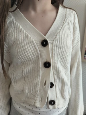 Vit kabelstickad kofta med knappar - Snygg vit kabelstickad kofta med stora mörka knappar framtill. Koftan har v-ringning, ribbade muddar och en croppad passform. Tillverkad i mjukt stickat material som känns skönt mot huden. Perfekt att slänga över en topp för en chill look. Passform är ganska stor.
