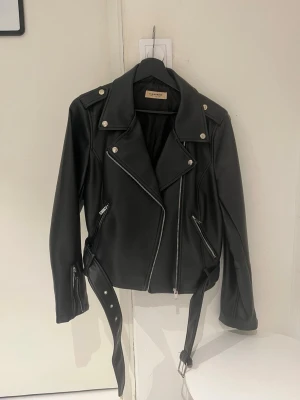 Svart skinnjacka från Glamorous - Snygg svart skinnjacka från Glamorous med klassisk bikerstil, silvriga dragkedjor och knappar samt bälte i midjan. Perfekt för dig som vill ha en tidlös och trendig jacka.