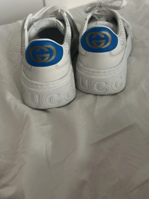 Gucci vita sneakers med blå detalj - Säljer ett par vita sneakers från Gucci med blå och gul logga på hälen samt klassiska röd-gröna ränder på sidan. Skorna har tjock platåsula, perforerade detaljer och är tillverkade i läder. Snygg och trendig modell som sticker ut.