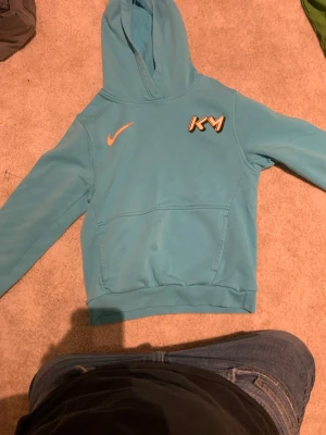 Turkos Nike hoodie med KM-logga - Snygg turkos hoodie från Nike med huva och magficka. På bröstet finns en orange Nike-logga och en KM-logga i vitt och orange. Hoodien har ribbade muddar vid ärmslut och nederkant. Perfekt för en avslappnad och sportig stil.