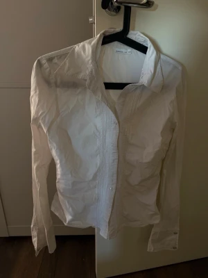 Vit skjorta från Stockh LM - Klassisk vit figursyddskjorta från Stockh LM med lång vidare ärm och knappar framtill. Skjortan har en snygg krage och diskreta sömdetaljer längs knappraden. Perfekt till både jeans och kostymbyxor för en clean look. 1 knapp saknas på ena ärmen (bild 5)
