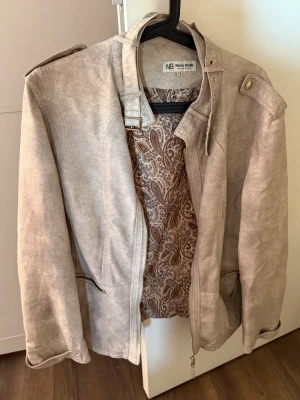 Beige skinnjacka från Nana Belle - Snygg beige skinnjacka från Nana Belle med coolt paisleymönstrat innerfoder. Jackan har axelklaffar, ståkrage med spänne och dragkedja framtill. Fickor med dragkedja och detaljer i metall ger en trendig look.