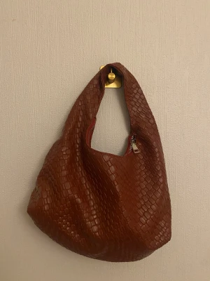 Röd  axelväska i skinn med mönster - Säljer en röd axelväska i skinn med snyggt, rutigt präglat mönster. Väskan har en mjuk, rundad form och stängs med dragkedja upptill. Perfekt storlek för att få plats med allt du behöver och har en cool, modern vibe.