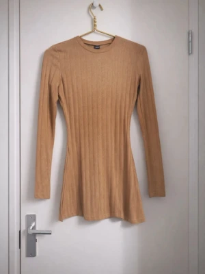 Beige ribbad klänning med lång ärm - Snygg beige ribbad klänning med lång ärm och rund hals. Klänningen har en figurnära passform och är tillverkad i ett mjukt material som ger en bekväm känsla. Perfekt för dig som gillar stilrena och enkla plagg.