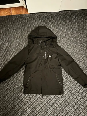 Svart Nike vindjacka med huva - Snygg svart vindjacka från Nike med huva och dragkedja framtill. Jackan har två fickor med dragkedja och meshfoder på insidan för extra komfort. Nike-logga på bröstet och justerbar huva för bästa passform. Perfekt för blåsiga dagar.