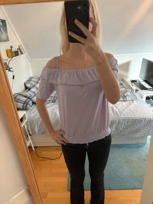 Ljuslila offshoulder topp - Säljer en ljuslila offshoulder topp med resår i nederkant och vid axlarna. Toppen har korta ärmar och en volangdetalj längs axelpartiet. Perfekt för dig som gillar en avslappnad men söt stil.
