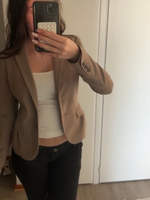 Beigebrun kavaj med knappdetaljer - Snygg beige kavaj med figurnära passform och klassisk krage. Kavajen har två framfickor, dekorativa knappar vid ärmslut och stängs med en knapp framtill. Perfekt för en stilren look och enkel att matcha med olika outfits.