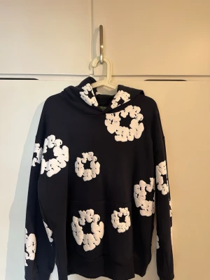Denim Tears - Snygg hoodie från Denim Tears med stora vita blommönster tryckta över hela tröjan. Hoodien har en klassisk huva och magficka framtill. Perfekt för dig som vill sticka ut med en unik och trendig stil.