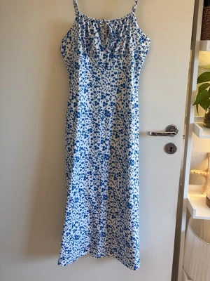 Blommig långklänning med smala band - Söt långklänning med smala axelband och blått blommönster på vit botten. Klänningen har en rak passform och liten knytning vid urringningen. Perfekt för varma dagar och somriga tillfällen.
