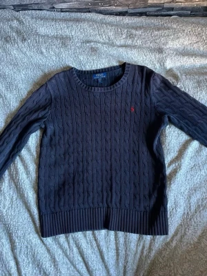 Mörkblå kabelstickad tröja Polo - Kabelstickad mörkblå tröja från Polo Ralph Lauren med rund halsringning och lång ärm. Tröjan har ett litet rött broderat logomärke på bröstet och ribbade muddar vid ärmslut och nederkant. Klassisk och tidlös design i mjukt material.
