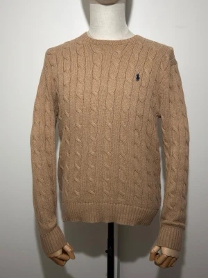 Beige kabelstickad tröja Ralph Lauren - Beige kabelstickad tröja från Polo Ralph Lauren med rund hals och mörkblå broderad logga på bröstet. Tröjan har långa ärmar och ribbade muddar vid ärmslut och nederkant. Klassisk modell i mjukt material. I nyskick utan defekt. Nypris ca 2000 kr.