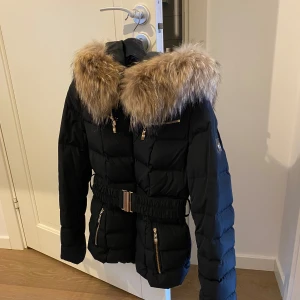 Svart dunjacka med pälsdetaljer från Hollies - Snygg svart dunjacka från Hollies med lyxig päls på huvan och guldfärgade dragkedjor. Jackan har ett bälte i midjan för en figursydd look och flera praktiska fickor. Perfekt för kyliga dagar.