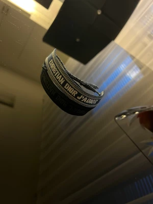 Grått tygarmband från Dior - Snyggt marinblått tygarmband från Christian Dior med texten 'J'ADIOR' invävd i vitt. Armbandet har justerbar knytning och cool bohemisk vibe. Perfekt accessoar för att lyfta din outfit med en exklusiv touch.