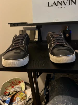 Svarta sneakers från Lanvin - Snygga svarta sneakers från Lanvin med vita sulor och svarta skosnören. Skorna har en låg profil och blanka partier på ovandelen som ger en clean look. Metallöljetter och diskret söm längs sidan. Perfekta för dig som gillar stilrena och exklusiva sneakers.(är öppen för byten)
