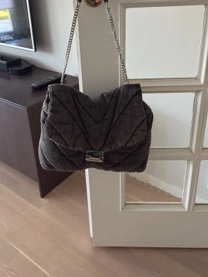 Mörkgrå quiltad handväska med kedja - Säljer en snygg mörkgrå quiltad handväska med silverfärgad kedjerem och metallspänne. Väskan har en mjuk, puffig design och är tillverkad i ett tyg som liknar manchester. Den är i väldigt bra skick då den knappt är använd!😇💗🌸