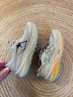 Beige Asics Trabuco Max 3 sneakers - Snygga beige Asics Trabuco Max 3 sneakers, jogging/trail-skor med mesh-överdel, svarta detaljer och turkos sula. Skorna har en tjock, dämpad sula med orange inslag och klassisk Asics-logga på sidan. Perfekta för dig som gillar sportig stil och komfort eller gillar att löpträna i tex skog. Mycket bra skick, endast använda en gång och säljes pga för små.