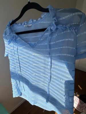 Ljusblå blus med volang från Happy Holly - Söt ljusblå blus från Happy Holly med transparenta ränder, volangdetaljer vid halsen och knytband framtill. Blusen har korta ärmar och v-ringning, perfekt för vår och sommar. Materialet känns lätt och luftigt🥂💗