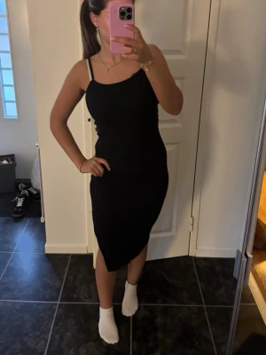 Svart långklänning med slits - Säljer en svart långklänning med smala axelband och slits på sidan. Klänningen har en figurnära passform och är perfekt för dig som gillar en enkel men stilren look. Materialet känns mjukt och stretchigt, vilket gör den bekväm att bära.