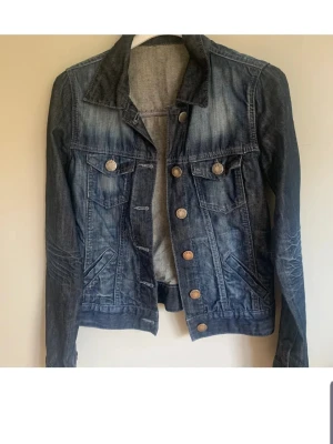 Mörkblå klassisk jeansjacka - Snygg mörkblå jeansjacka med slitningar och kontrastsömmar. Jackan har två bröstfickor med knappar, sidofickor och metallknappar framtill. Tidlös modell med krage och justerbar nederkant. Perfekt att slänga över en hoodie eller t-shirt.