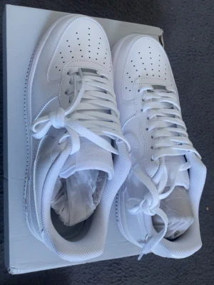 Nike Air Force 1 vita sneakers - Klassiska Nike Air Force 1 sneakers i helvitt läder med perforerad tåbox, vita skosnören och den ikoniska swoosh-loggan på sidan. Låg modell med robust sula och diskret branding på plösen och hälen. Perfekta för en clean och stilren look.