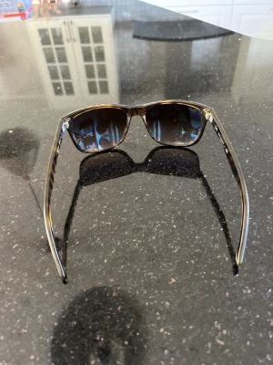 Bruna Ray-Ban solglasögon - Snygga solglasögon från Ray-Ban med bruna bågar och mörka glas. Klassisk fyrkantig form och tillverkade i Italien. Perfekta för soliga dagar och ger en cool vibe till din outfit.