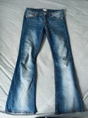 Blå lågmidjade bootcut jeans  - Säljer ett par blå bootcut jeans med snygga slitningar och broderade bakfickor. Jeansen har klassisk femficksmodell, låg midja och är tillverkade i stretchigt denimtyg för skön passform. Se defekter på bilderna 