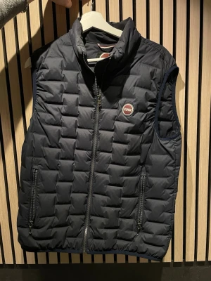 Marinblå dunväst från Colmar - Snygg marinblå dunväst från Colmar med quiltat mönster och hög krage. Västen har två dragkedjeförsedda fickor och ett rundat Colmar-märke på bröstet. Perfekt att slänga över en hoodie eller tröja för extra värme och stil. Pris går att diskutera