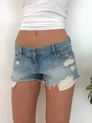 Hollister lågmidjade slitna jeansshorts  - Ett par jeansshorts från Hollister i storlek W27, vilket motsvarar en vanlig S. Rakt över midjemått: ca 38cm. Observera, modellen brukar ha XXS, därav svårt att visa förväntad passform för S. Jag fraktar inom 24h, frakt med PostNord prioriteras. Se liknande plagg i min profil!