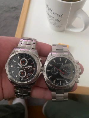 Casio Edifice och Gant herrklockor - Två stilrena herrklockor: Casio Edifice med svart urtavla, orange detaljer och kronograffunktion samt Gant med svart urtavla, datumvisning och tachymeter. Båda har robusta armband i silverfärgat stål och sportig design.      Behöver batteri byta 