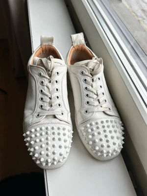 Vita sneakers med nitar från Louboutin - Unika vita sneakers från Christian Louboutin med coola vita nitar över hela tån. Skorna har klassisk låg profil, vita snören och detaljer i läder. Insidan är beige och designen är riktigt edgy för dig som vill sticka ut.