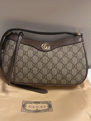 Gucci GG canvas axelremsväska brun - Snygg axelremsväska från Gucci i klassiskt GG canvasmönster med bruna läderdetaljer och guldfärgad logga framtill. Väskan har en avlång form och justerbar rem, perfekt för att bära över axeln. Kommer med dustbag.